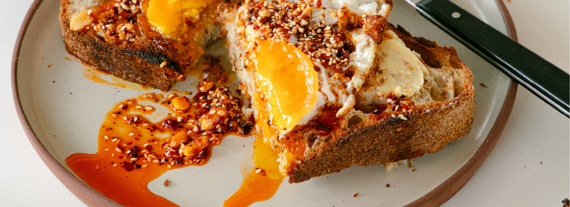A slice of toast cut in half on a white plate with butter, eggs and White Mausu peanut rāyu on top. A piece of cutlery with a black base is half visible on the plate
