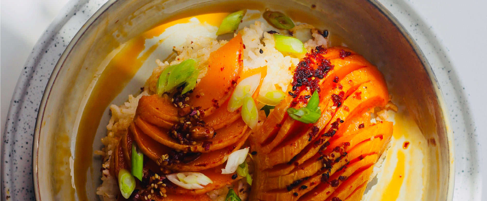 A off white bowl on a speckled plate containing sliced sweet potato, spring onions and rice topped with White Mausu peanut rāyu