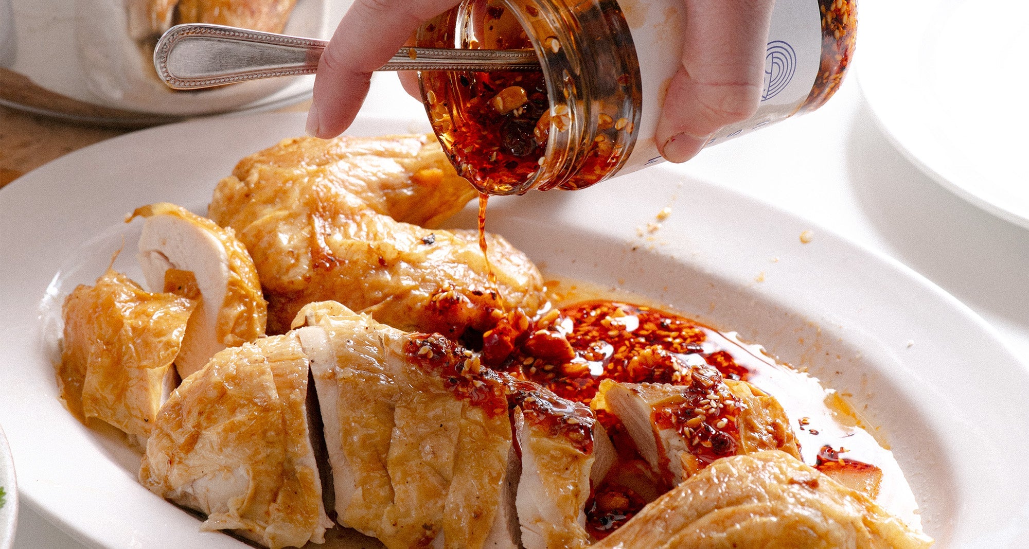 cooked chicken on a white plate with a open jar of White Mausu Peanut Rāyu being poured over with a silver spoon in it