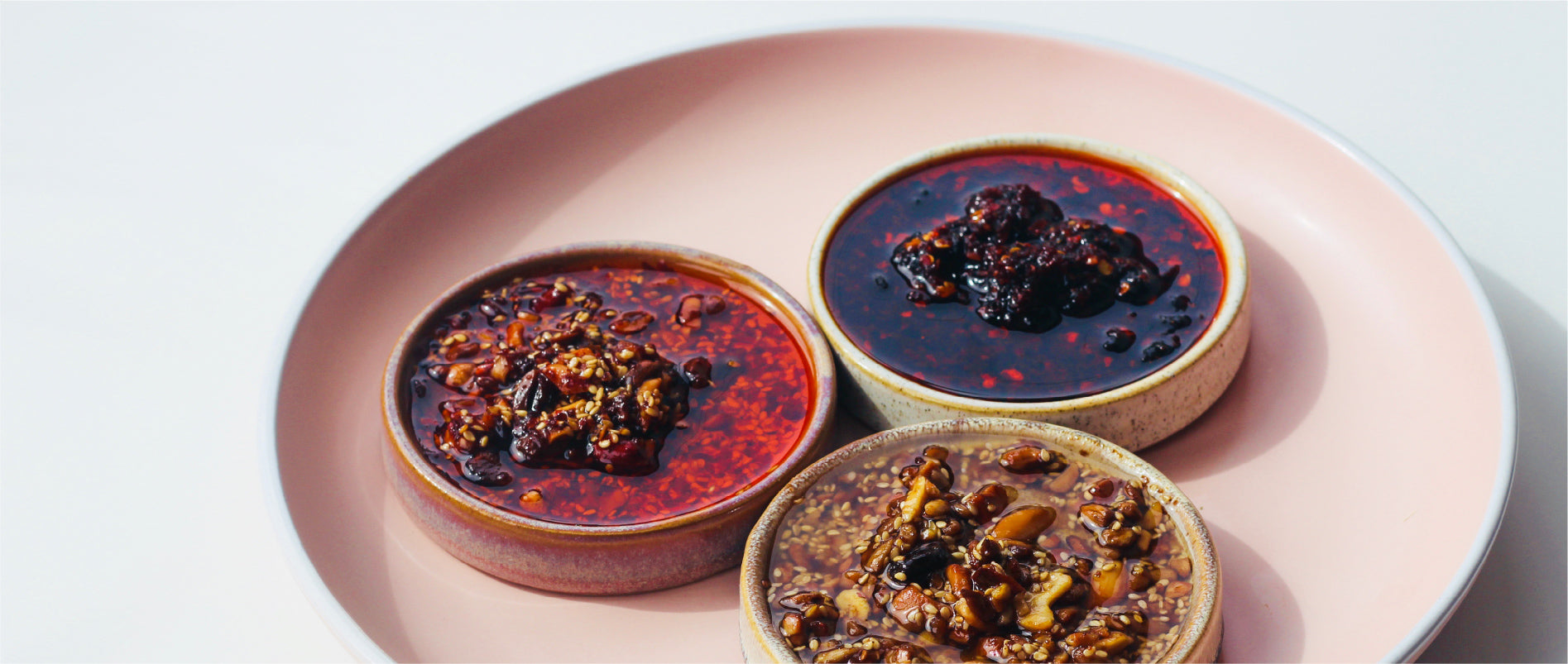 three chilli oils, peanut rāyu, black bean and smoky chilli oil in ceramic bowls on a pink plate.