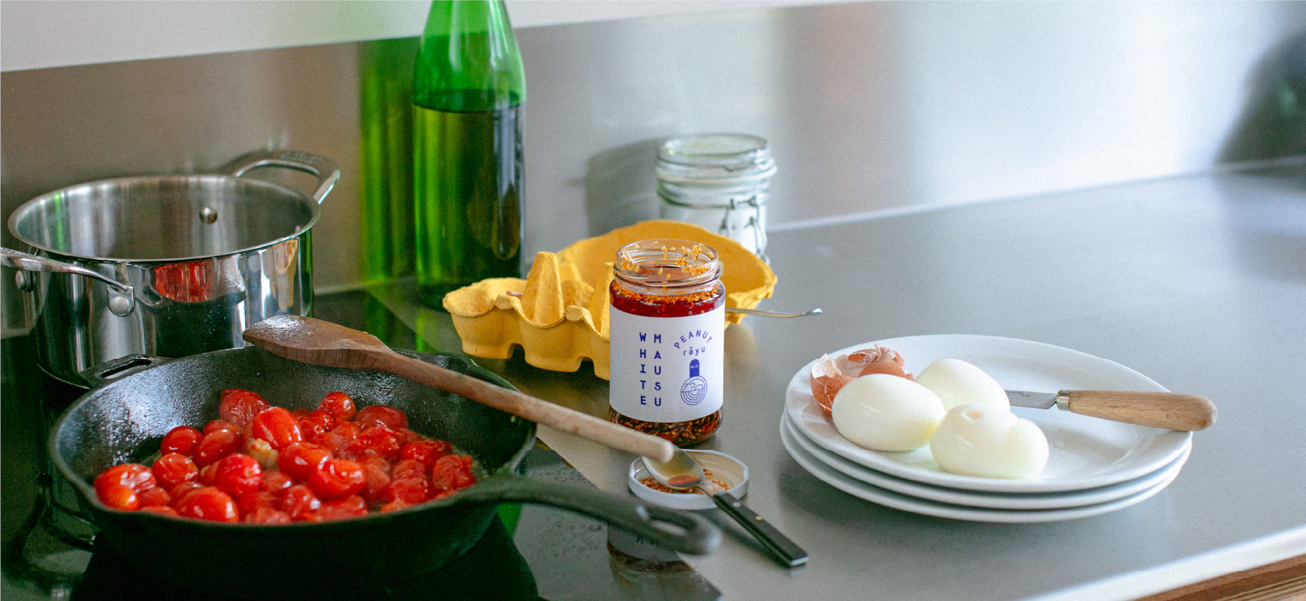 Kitchen countertop with cherry tomatoes in a cast iron pan and a wooden spoon lying on top, a silver cooking pot behind it. To the right are a green glass bottle, a glass container with a white substance, an empty egg carton, an open jar of White Mausu peanut rāyu, a teaspoon sitting on the lid and a stack of three white plates with three hard boiled eggs, broken egg shells and a knife
