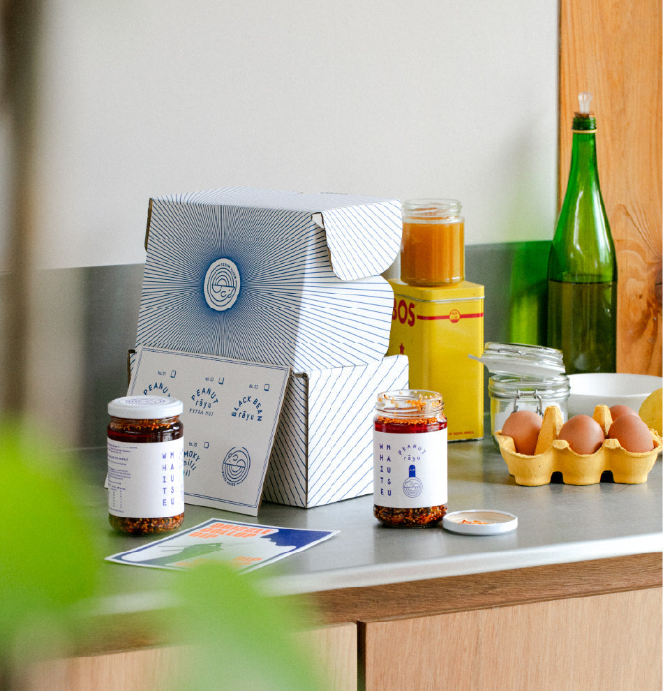 An assortment of food and jars on a counter top which includes 2 jars of White Mausu peanut rāyu, 2 postcards, a carton of eggs, a white box with the white mausu logo and design in blue, a green glass bottle, a glass jar, a white bowl, a jar filled with orange substance and a yellow tin box with red writing