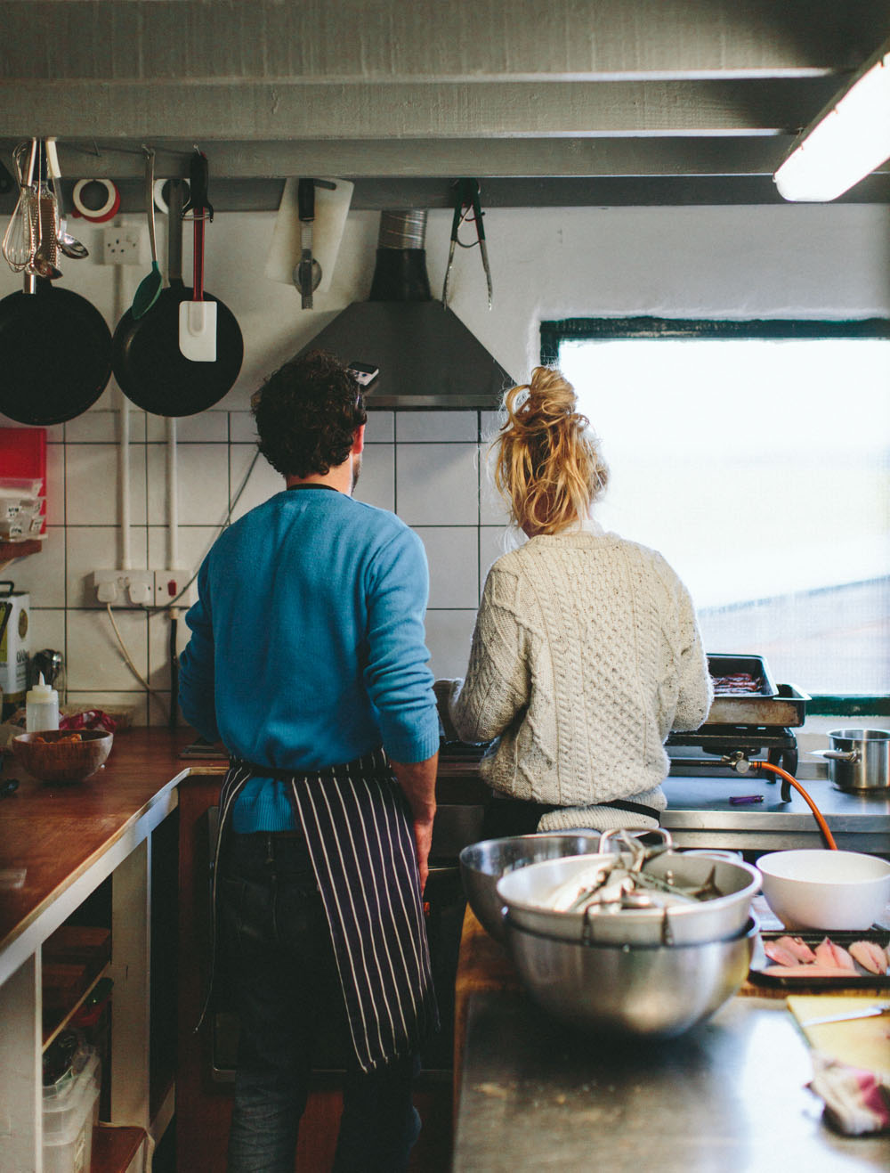 Two people standing in a kitchen facing away from the camera. One is wearing a blue jumper, jeans and a blue apron. The other is wearing a white knitted jumper with their hair tied up in a bun. Various kitchen items sit on the countertop with two pans hanging from the ceiling.